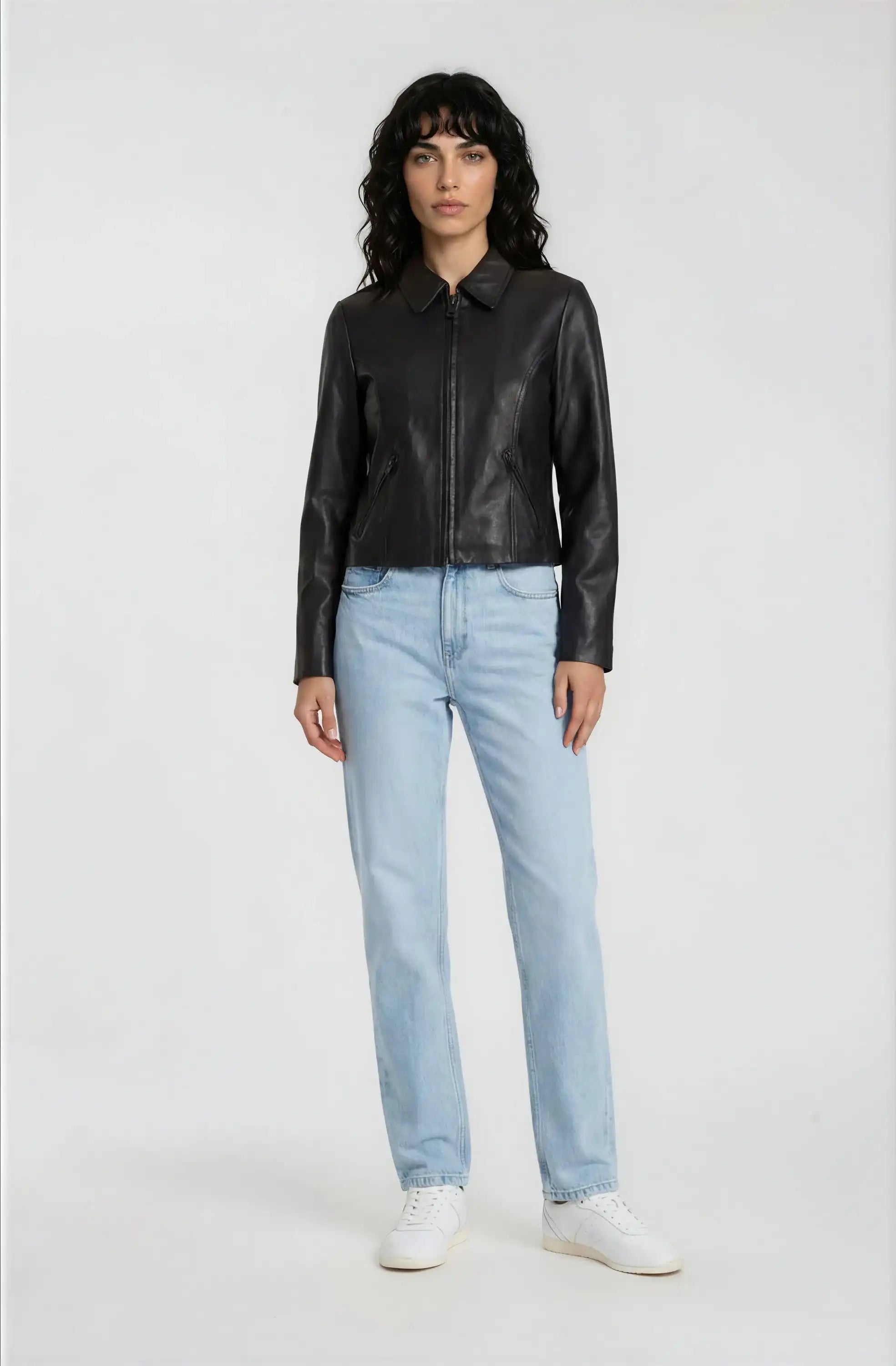 Eine Frau trägt die trueprodigy Damen Lederjacke aus Schafsleder in Schwarz mit minimalistischem Design, kombiniert mit hellblauen Jeans und weißen Sneakern, vor schlicht weißem Hintergrund.