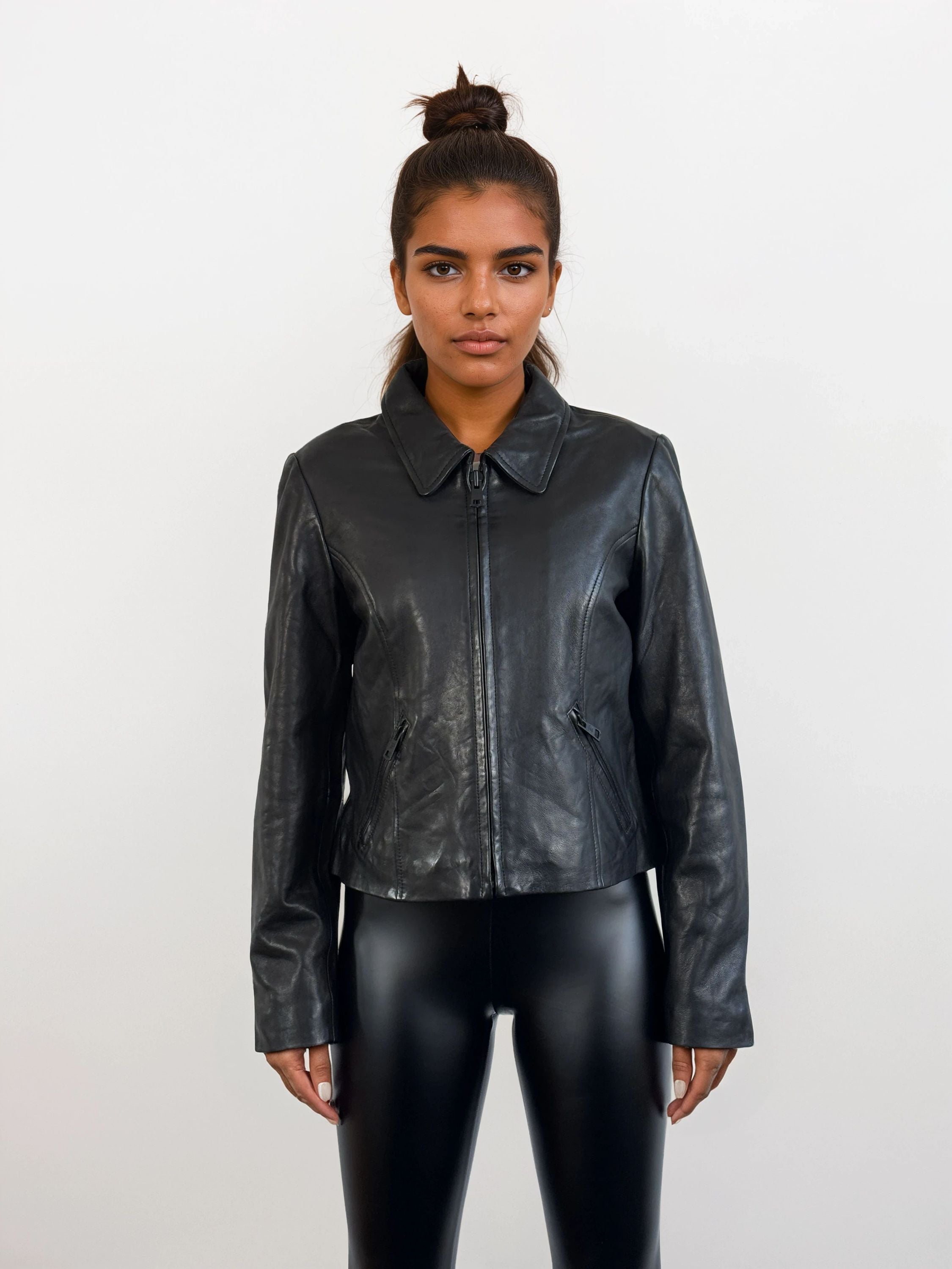 Eine Person steht mit dem Gesicht nach vorne vor einem weißen Hintergrund und trägt die trueprodigy Damen Lederjacke aus Schafsleder in Schwarz mit minimalistischem Design und schwarz glänzenden Leggings. Ihr Haar ist zu einem Dutt hochgesteckt, die Arme ruhen natürlich an der Seite.