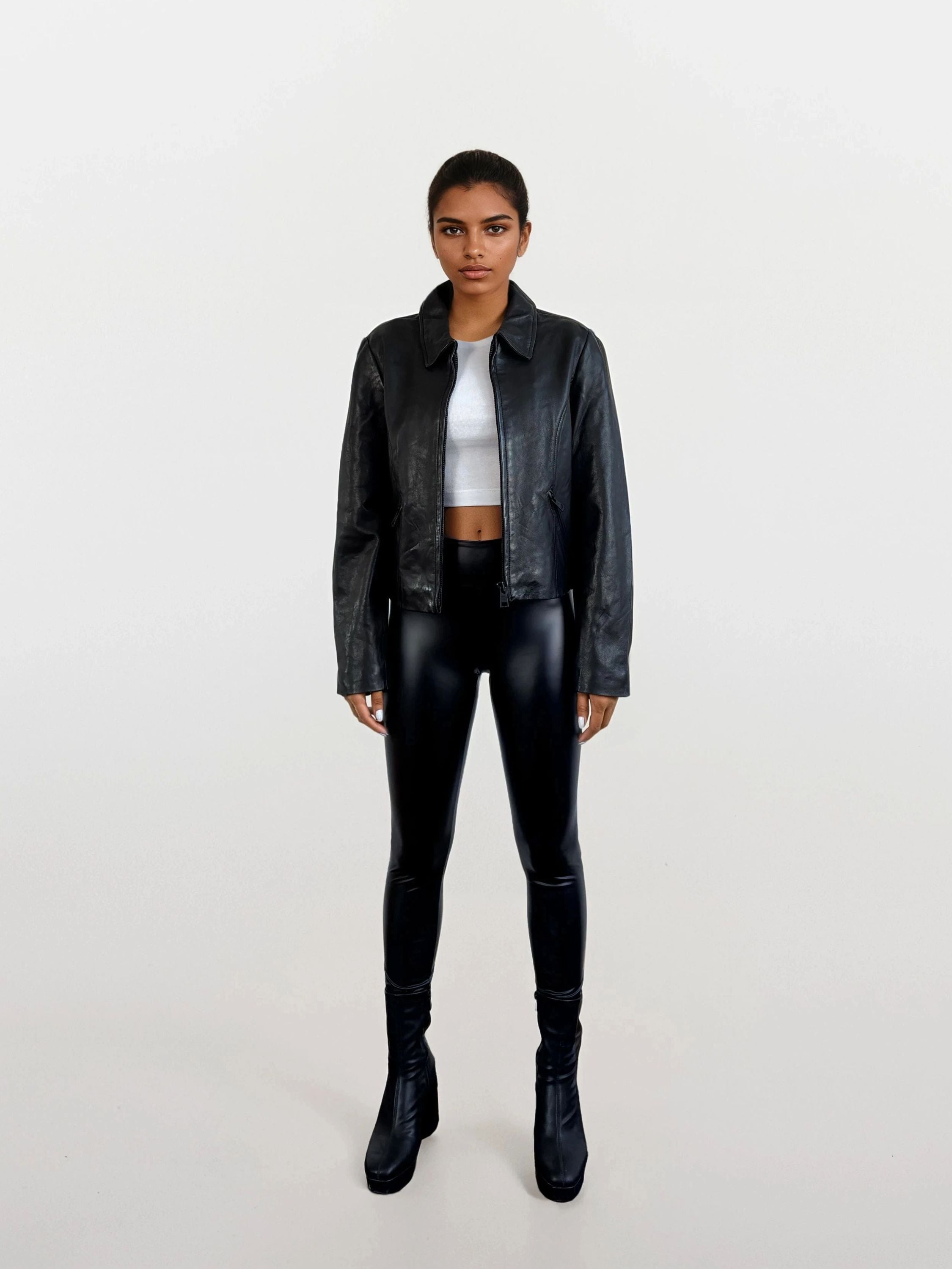 Eine Person steht mit dem Gesicht nach vorne vor einem schlichten weißen Hintergrund und trägt die schwarze Damen Lederjacke aus Schafsleder mit minimalistischem Design von trueprodigy, schwarze glänzende Leggings, ein weißes Crop-Top und schwarze hochhackige Stiefel.
