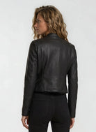 Eine Person mit gewelltem, hellbraunem Haar trägt die trueprodigy Damen Lederjacke aus Schafsleder in Schwarz im sportlichen Racing-Stil und eine schwarze Hose und blickt von der Kamera weg auf einen schlichten, hellen Hintergrund.