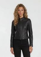 Eine Person mit gewelltem, hellbraunem Haar steht vor einem schlichten Hintergrund und trägt die trueprodigy Damen Lederjacke aus Schafsleder in Schwarz im sportlichen Racing-Stil, gepaart mit einer schwarzen Hose.