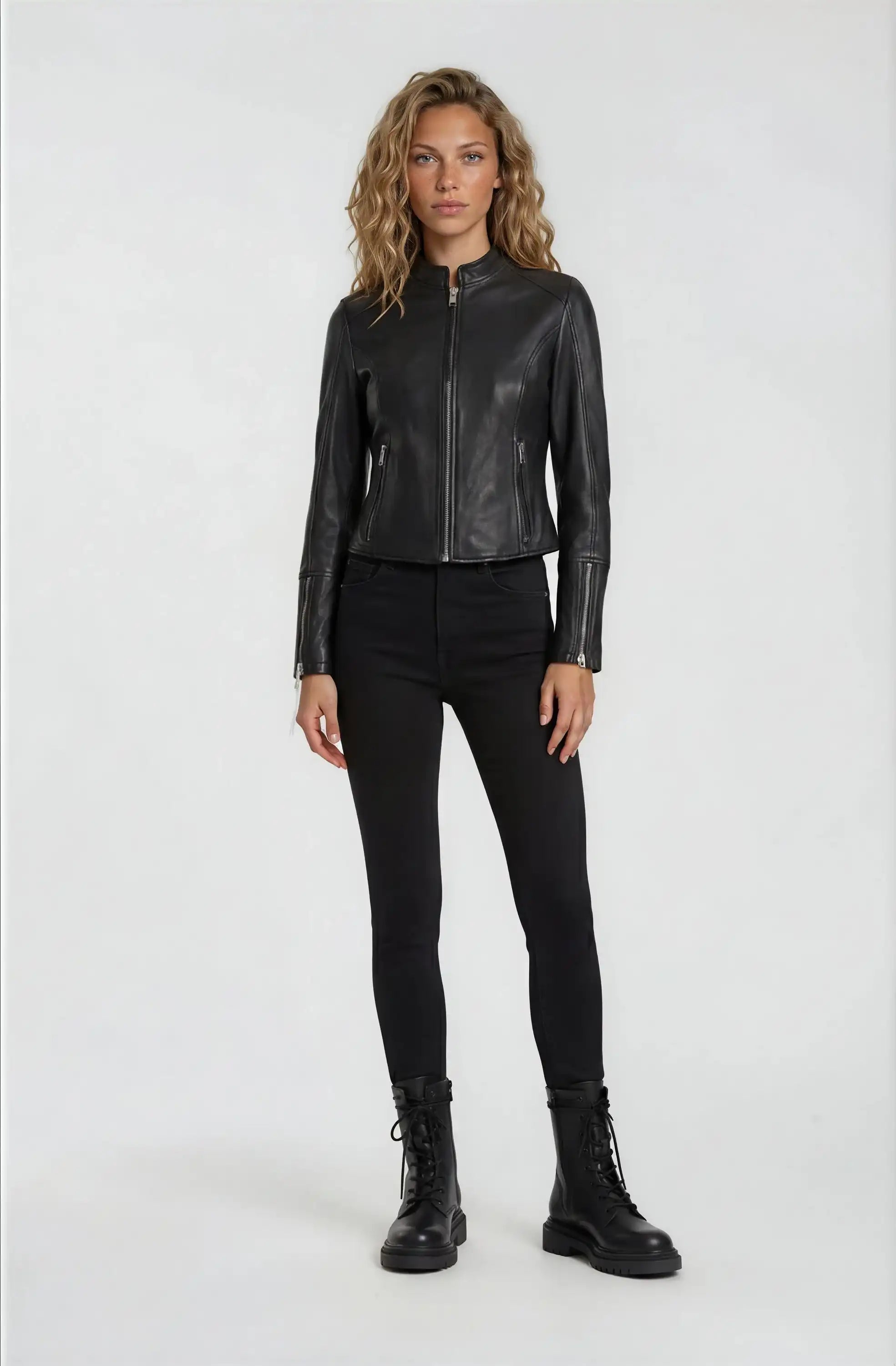 Eine Person sieht nach vorne und trägt eine trueprodigy Damen Lederjacke aus Schafsleder in Schwarz im sportlichen Racing-Stil, gepaart mit einer schwarzen Hose und Schnürstiefeln. Der Hintergrund ist einfarbig und hell.