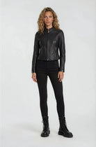 Eine Person sieht nach vorne und trägt eine trueprodigy Damen Lederjacke aus Schafsleder in Schwarz im sportlichen Racing-Stil, gepaart mit einer schwarzen Hose und Schnürstiefeln. Der Hintergrund ist einfarbig und hell.