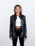 Eine Frau steht mit dem Gesicht nach vorne und trägt die trueprodigy Damen Lederjacke aus Schafsleder in Schwarz im sportlichen Racing-Stil über einem weißen Oberteil und einer schwarzen, glänzenden Hose mit hoher Taille, vor einem schlichten, hellen Hintergrund.