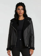 Eine Frau mit langen dunklen Haaren modelliert die trueprodigy Damen Lederjacke aus Schafsleder in Schwarz mit Teddy-Besatz über einem schwarzen Oberteil und einer Hose vor einem schlichten hellgrauen Hintergrund.