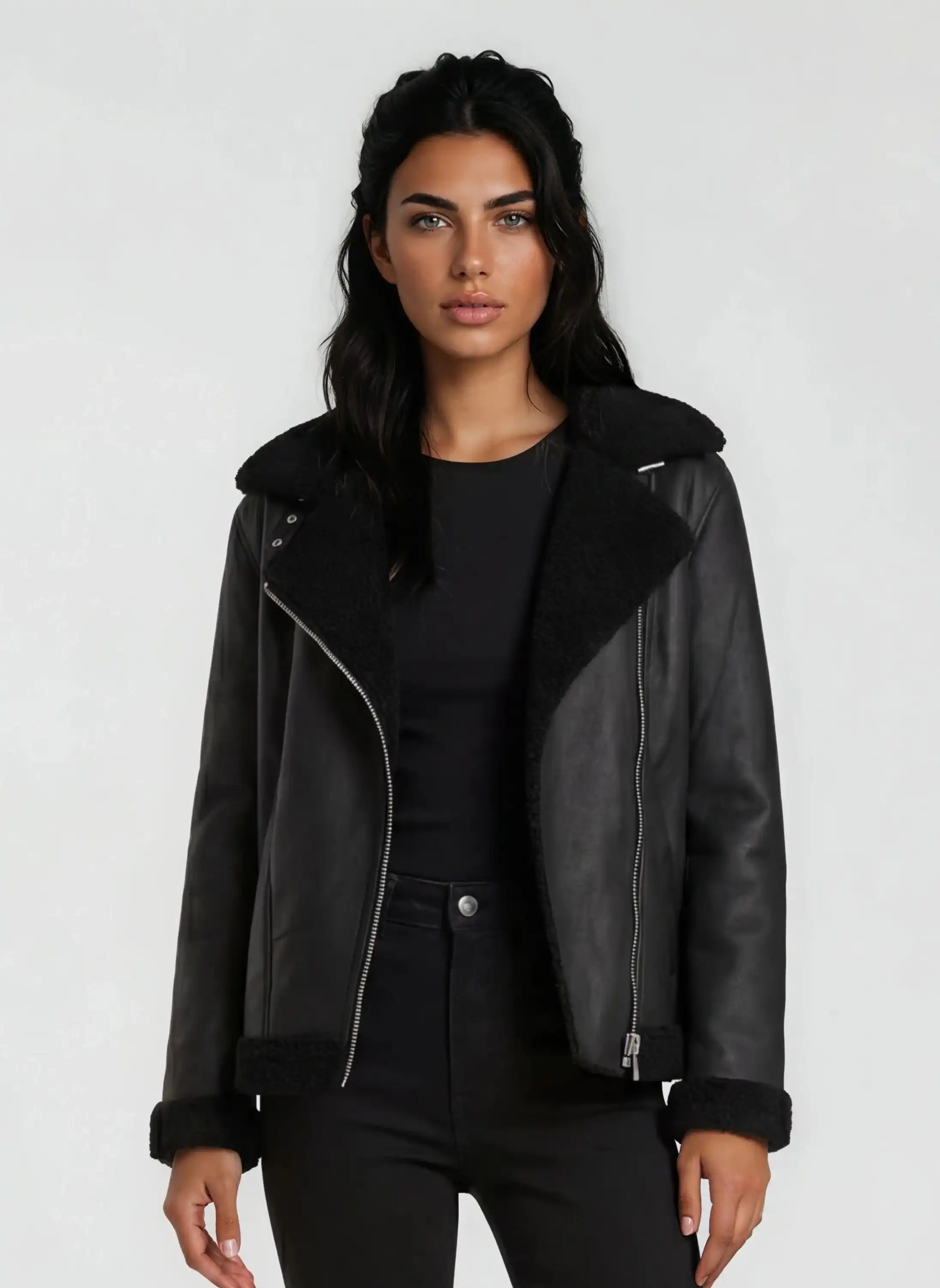 Eine Frau mit langen dunklen Haaren modelliert die trueprodigy Damen Lederjacke aus Schafsleder in Schwarz mit Teddy-Besatz über einem schwarzen Oberteil und einer Hose vor einem schlichten hellgrauen Hintergrund.
