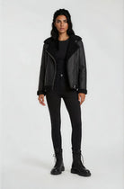 Eine Frau steht mit dem Gesicht nach vorne vor einem hellen Hintergrund und trägt die trueprodigy Damen Lederjacke aus Schafsleder in Schwarz mit Teddy-Besatz, gepaart mit einem schwarzen Top, Skinny Jeans und Schnürstiefeln. Ihre Arme ruhen an den Seiten.