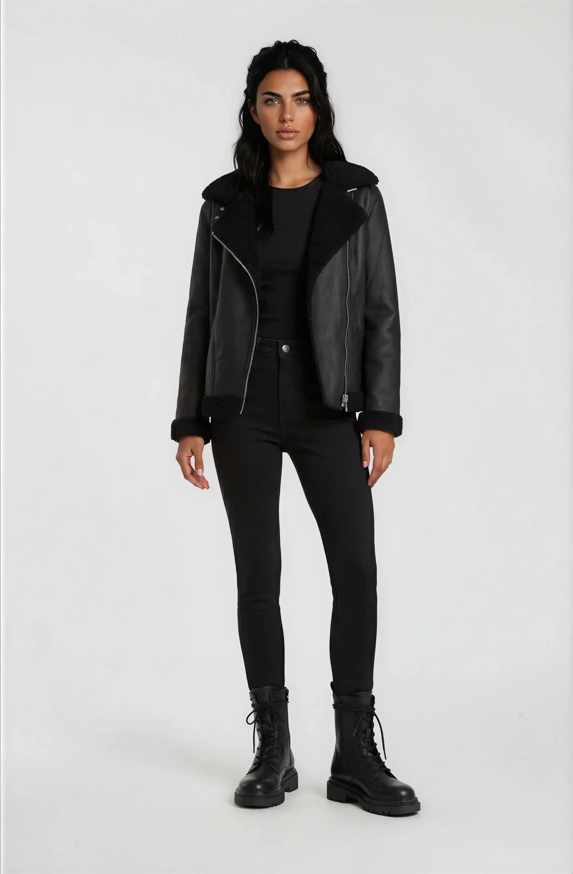 Eine Frau steht mit dem Gesicht nach vorne vor einem hellen Hintergrund und trägt die trueprodigy Damen Lederjacke aus Schafsleder in Schwarz mit Teddy-Besatz, gepaart mit einem schwarzen Top, Skinny Jeans und Schnürstiefeln. Ihre Arme ruhen an den Seiten.
