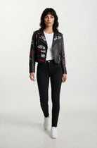 Eine Frau mit schulterlangen dunklen Haaren trägt die trueprodigy Feminine Lederjacke mit Grafittiprint und Reißverschluss - eine ausgefallene schwarze Lederjacke mit weißer und roter Grafik - mit einem weißen T-Shirt, schwarzen Jeans und weißen Turnschuhen und geht auf die Kamera zu.