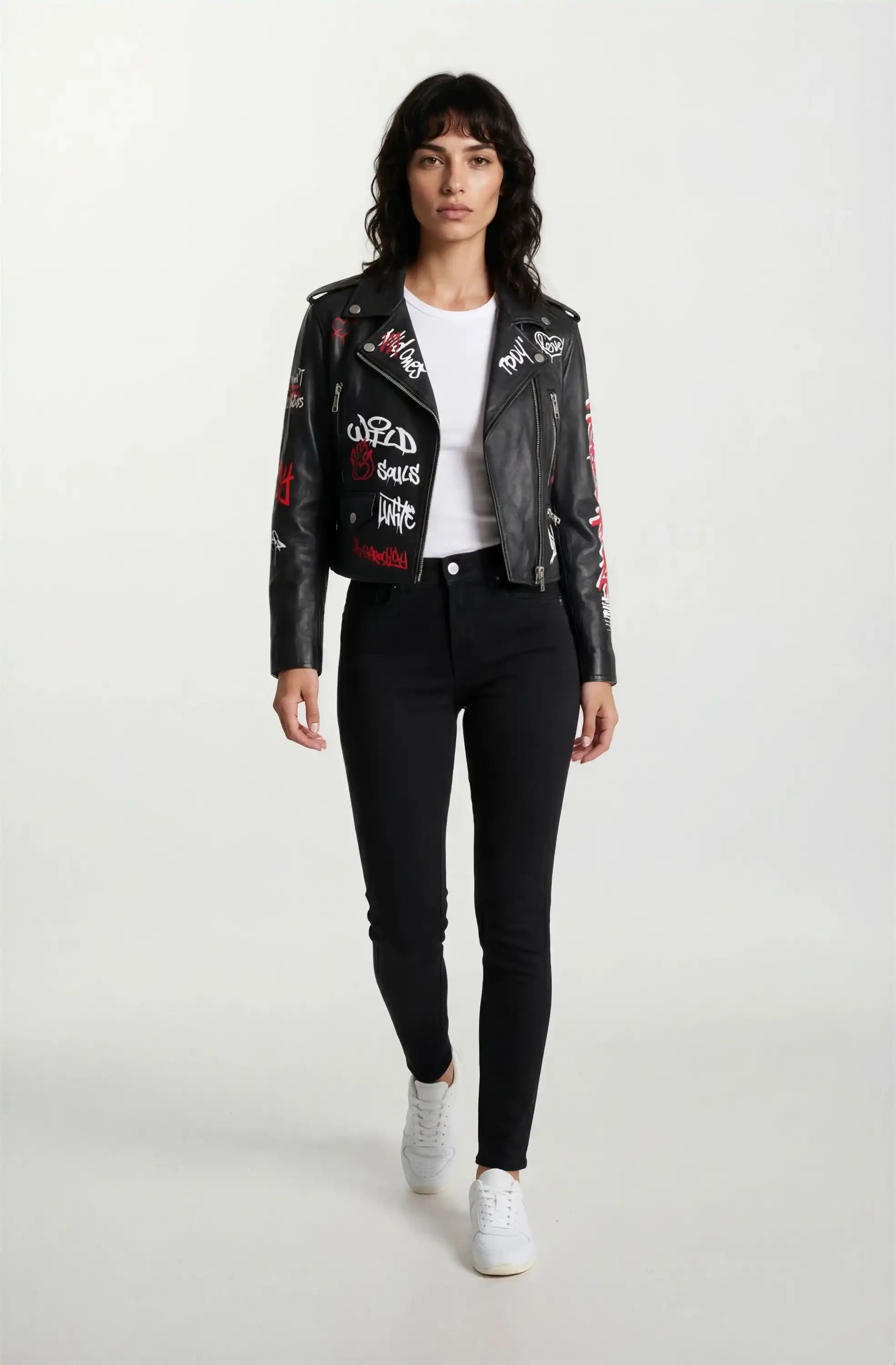 Eine Frau mit schulterlangen dunklen Haaren trägt die trueprodigy Feminine Lederjacke mit Grafittiprint und Reißverschluss - eine ausgefallene schwarze Lederjacke mit weißer und roter Grafik - mit einem weißen T-Shirt, schwarzen Jeans und weißen Turnschuhen und geht auf die Kamera zu.