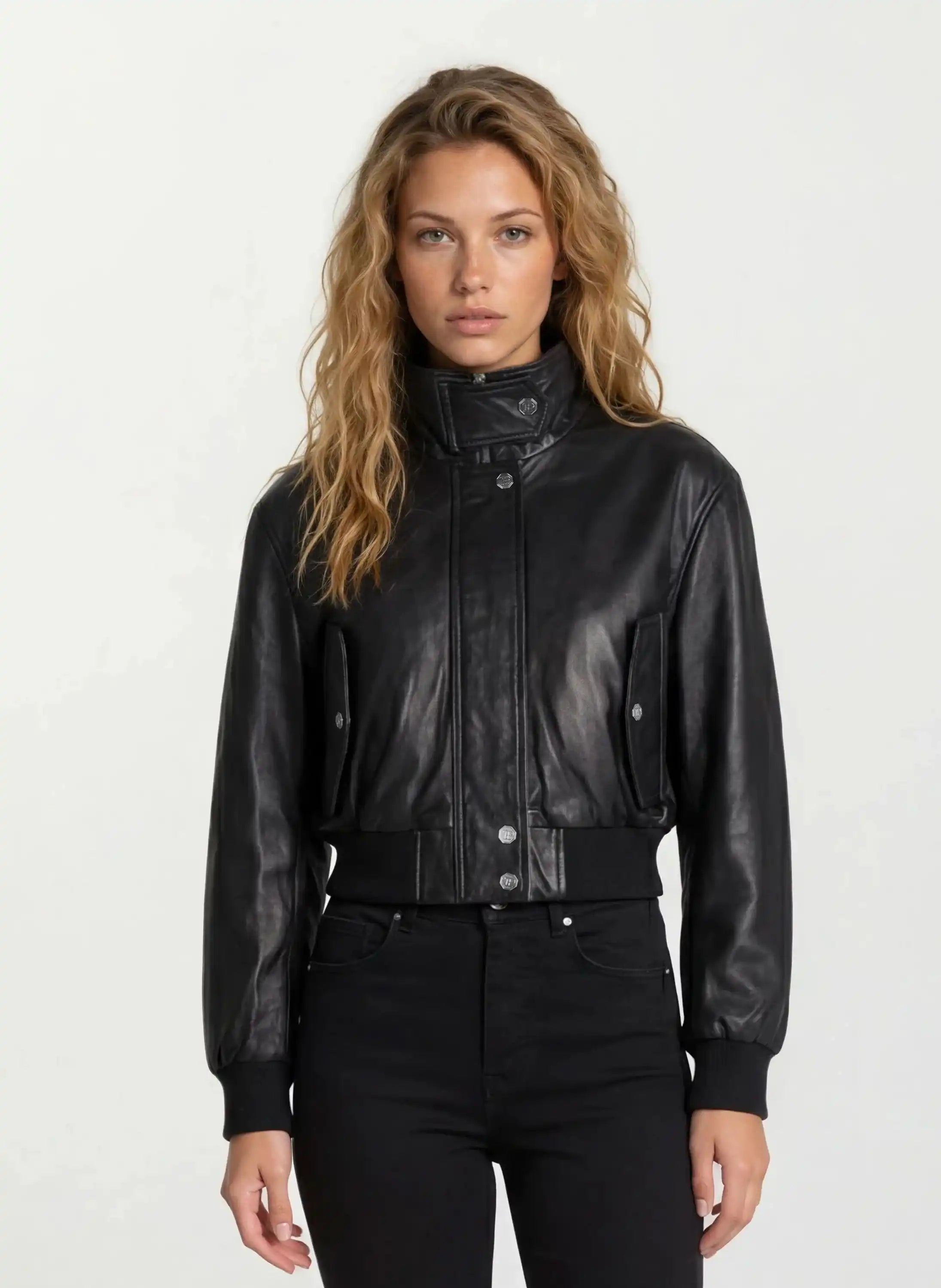 Eine Frau mit gewelltem, hellbraunem Haar trägt die trueprodigy Feminine Lederjacke Stehkragen und Reißverschluss und eine schwarze Hose und steht vor einem schlichten, hellen Hintergrund.