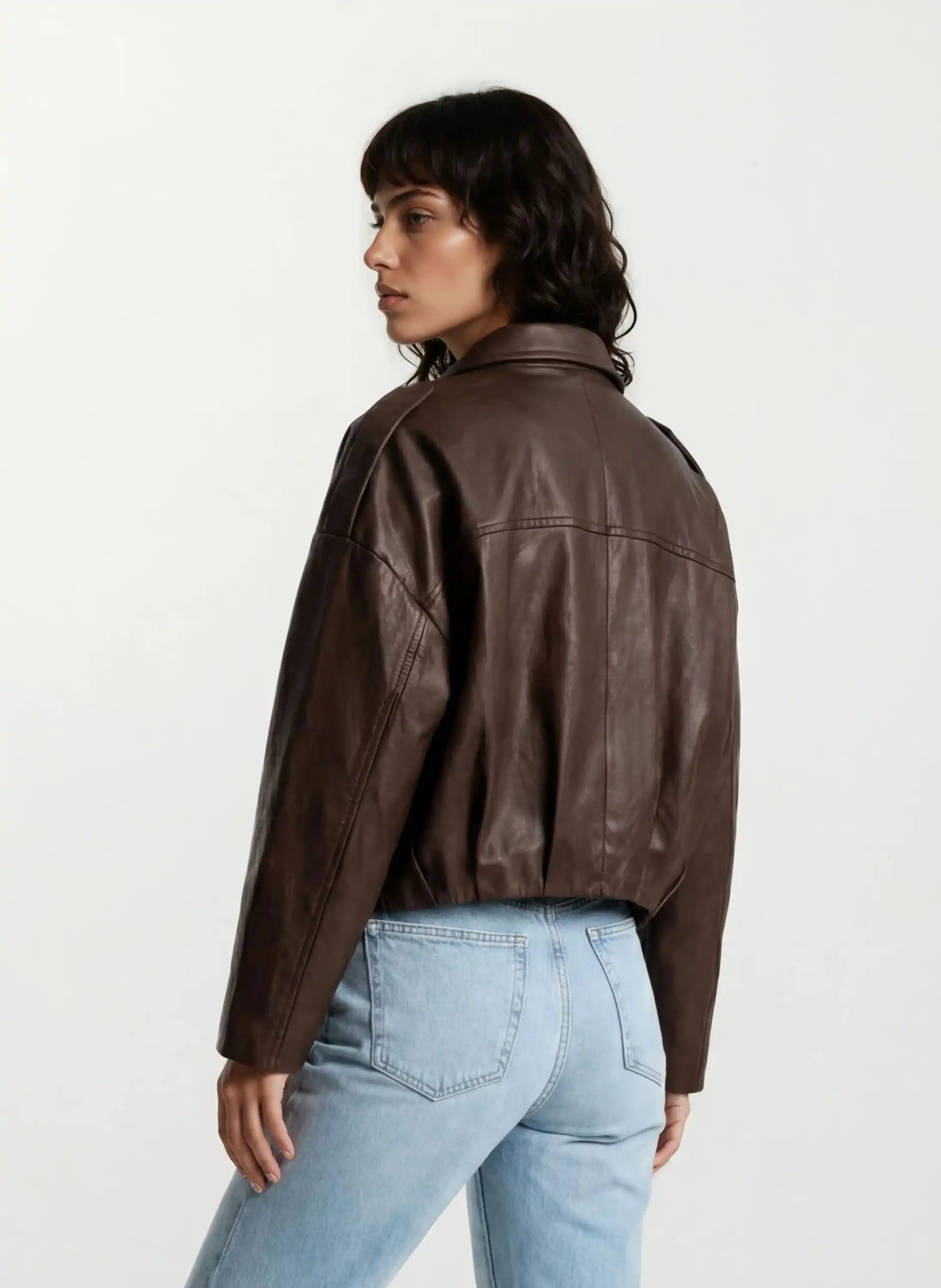 Eine Person mit gewellten dunklen Haaren trägt die trueprodigy Feminine Lederjacke mit Hemdkragen und Raffung am Saum und hellblaue Jeans und steht mit leicht gedrehtem Rücken und Kopf vor einem schlichten hellen Hintergrund.