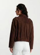 Eine Frau mit dunklem gewelltem Haar, von hinten gesehen, trägt die trueprodigy Feminine Lederjacke mit Hemdkragen und Raffung am Saum in brauner und weißer Hose vor einem schlichten, hellen Hintergrund.