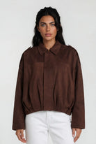 Eine Frau mit langen dunklen Haaren trägt die trueprodigy Clean-Cut Wildlederjacke Damen "Naeko" mit Hemdkragen und weißer Hose, steht vor einem schlichten hellen Hintergrund und blickt mit neutralem Gesichtsausdruck nach vorne.