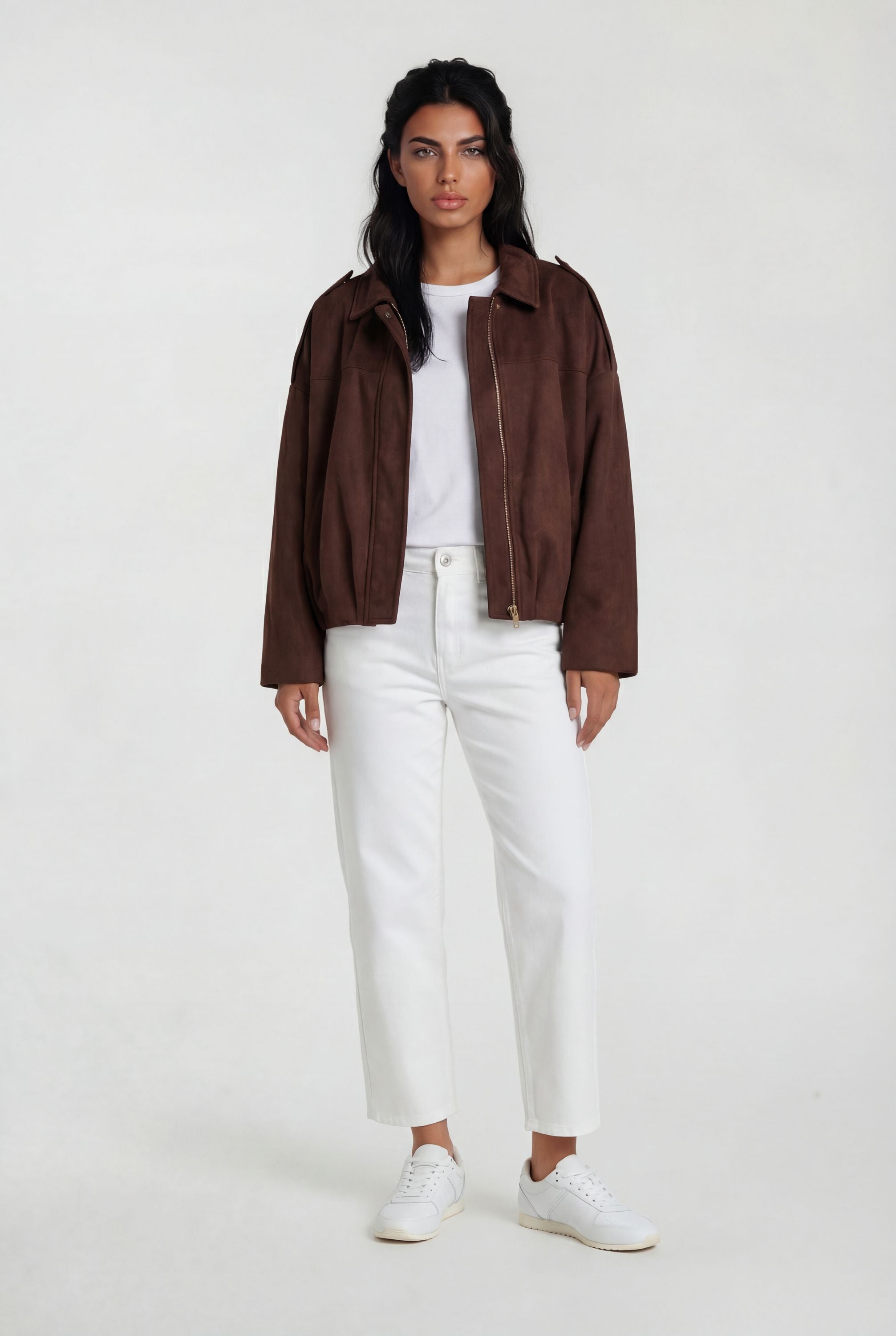 Eine Person steht mit dem Gesicht nach vorne und trägt die trueprodigy Clean-Cut Wildlederjacke Damen "Naeko" mit Hemdkragen, ein weißes T-Shirt, eine weiße Hose mit hoher Taille und weiße Turnschuhe vor einem schlichten hellen Hintergrund.