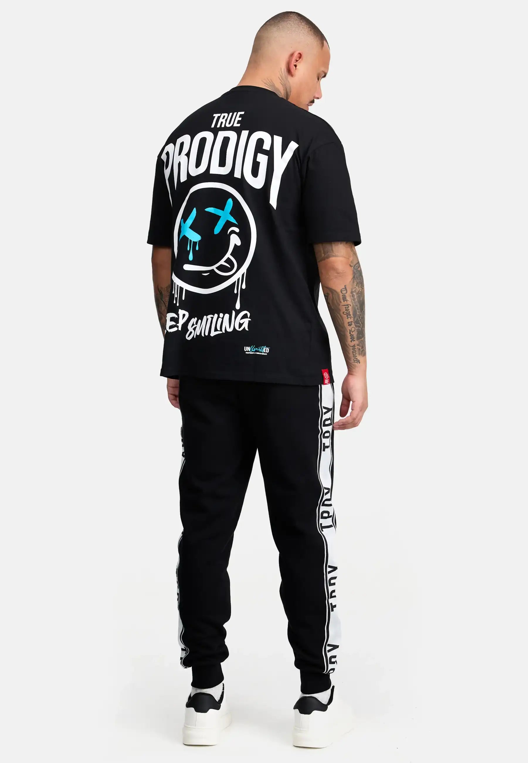Eine Person steht mit dem Gesicht zur Wand und trägt ein schwarzes TRUE PRODIGY T-Shirt mit großer Grafik auf dem Rücken, trueprodigy Weston Sweathose mit Logoschrift in schwarz mit weißen Streifen und weiße Turnschuhe. Der Hintergrund ist einfarbig hellgrau.