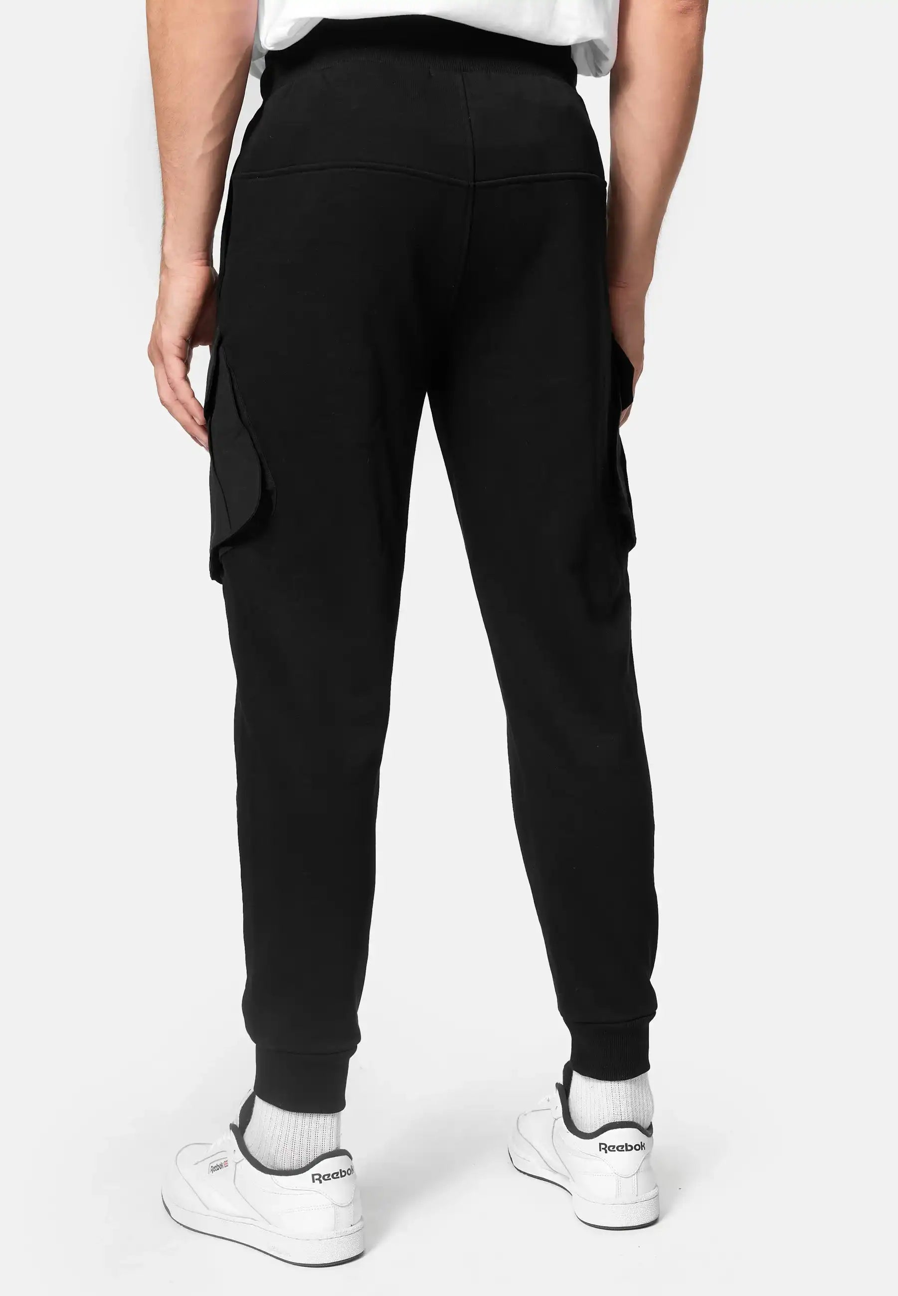 Eine Person steht von der Kamera abgewandt und trägt eine schwarze trueprodigy Stan Sweathose mit Cargotaschen, ein weißes T-Shirt, weiße Socken und weiße Reebok-Turnschuhe vor einem unifarbenen Hintergrund.