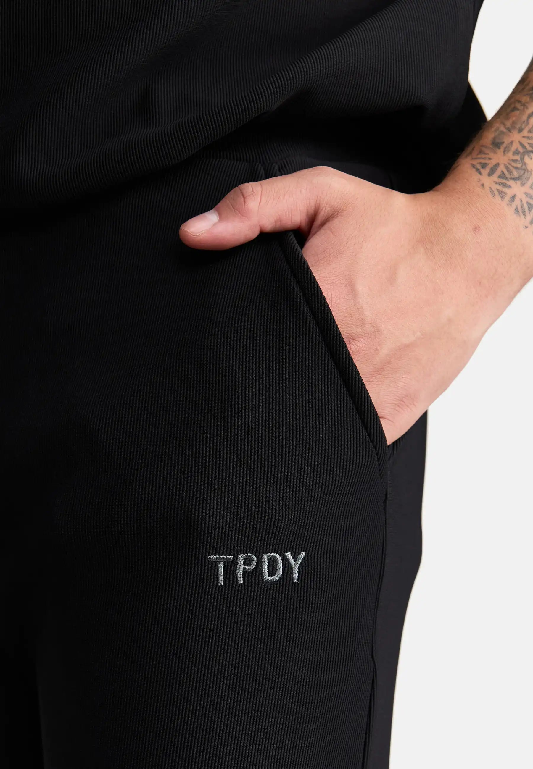 Eine tätowierte Person steckt ihre rechte Hand in die Tasche der schwarzen trueprodigy Ewan Jogger mit Stickerei, die in der Nähe der Tasche eine graue TPDY-Stickerei aufweist.