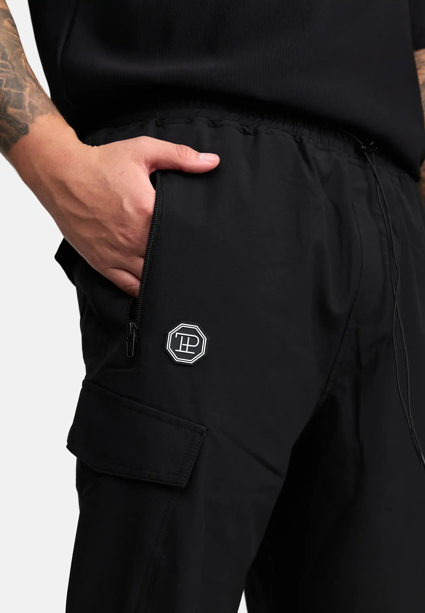 Eine Person in trueprodigy Jonny Leichte Cargo-Trainingshose mit Reißverschlusstasche und sechseckigem Logoaufnäher am Oberschenkel steht mit einer Hand in der Tasche. Auf ihrem Arm ist eine Tätowierung zu sehen.