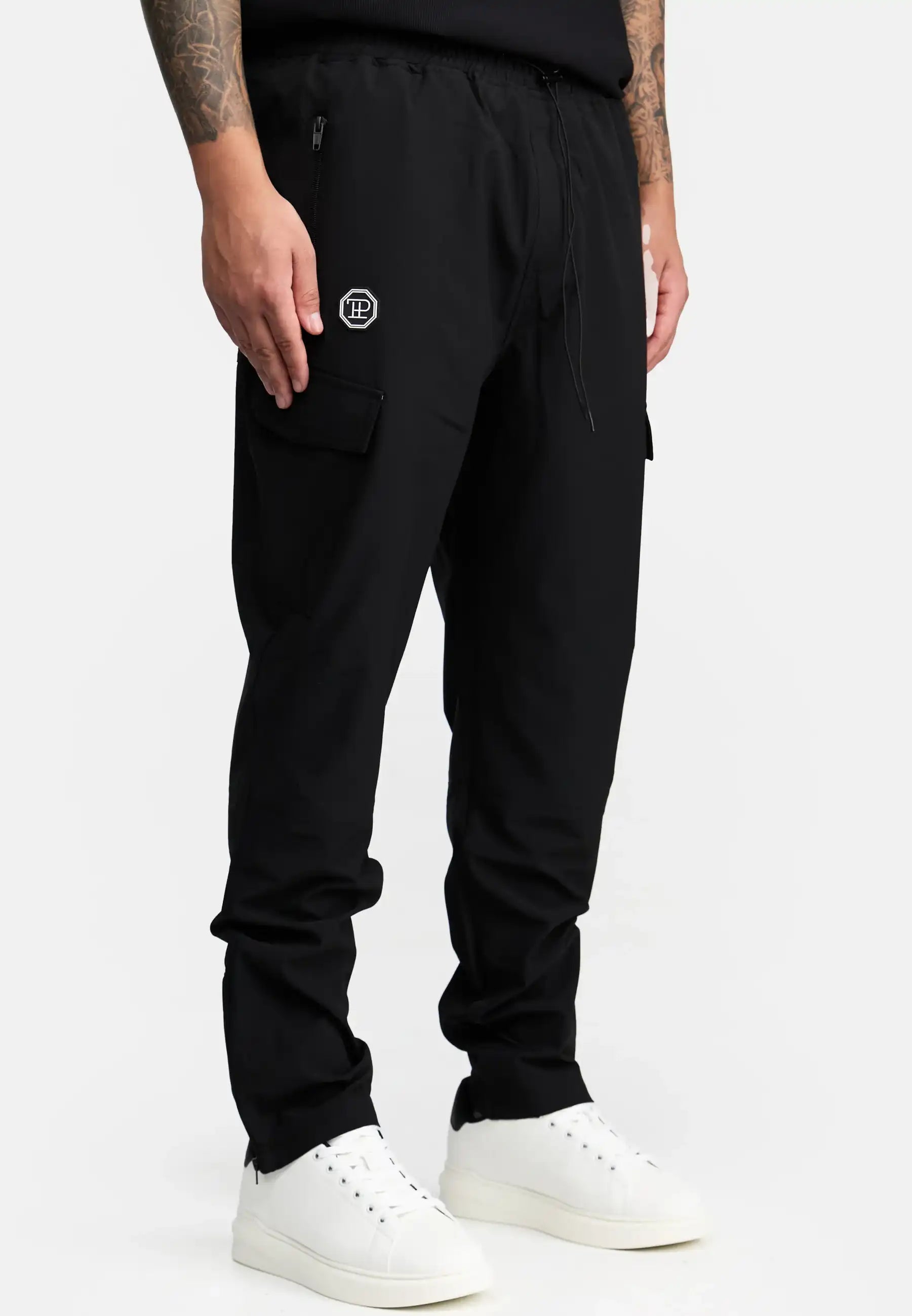 Eine Person trägt eine trueprodigy Jonny Leichte Cargo-Trainingshose in schwarz mit kleinem Logo auf dem linken Oberschenkel und Reißverschlusstaschen, gepaart mit einem schwarzen Hemd und weißen Sneakers. Der Hintergrund ist einfarbig weiß.