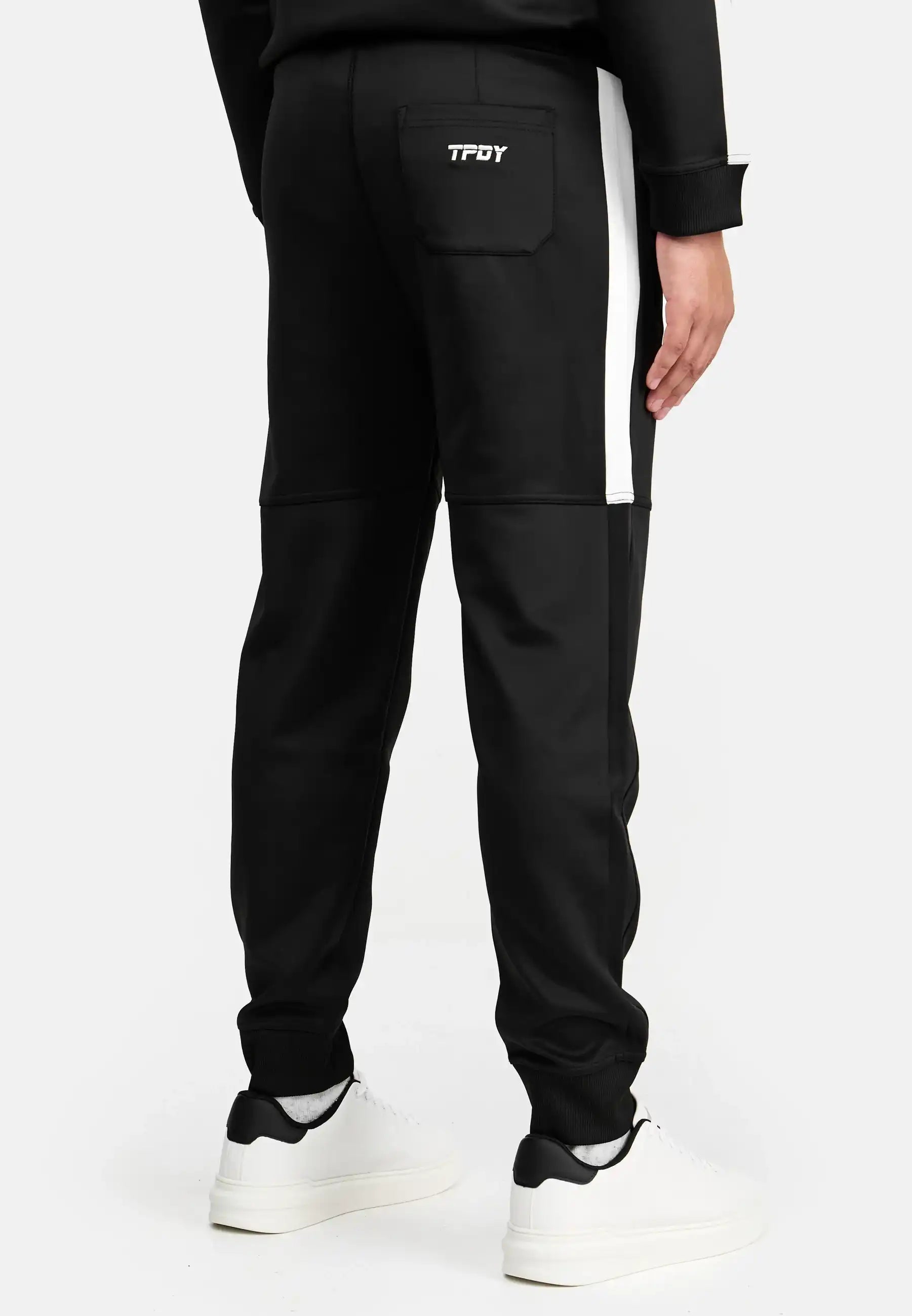 Abgebildet ist eine Person, die von der Taille abwärts die trueprodigy Francis Trainingshose mit Stickerei mit weißem Kontraststreifen trägt, gepaart mit weißen Sneakers. Die Hose hat eine kleine Gesäßtasche mit aufgesticktem "TFDY" darüber.