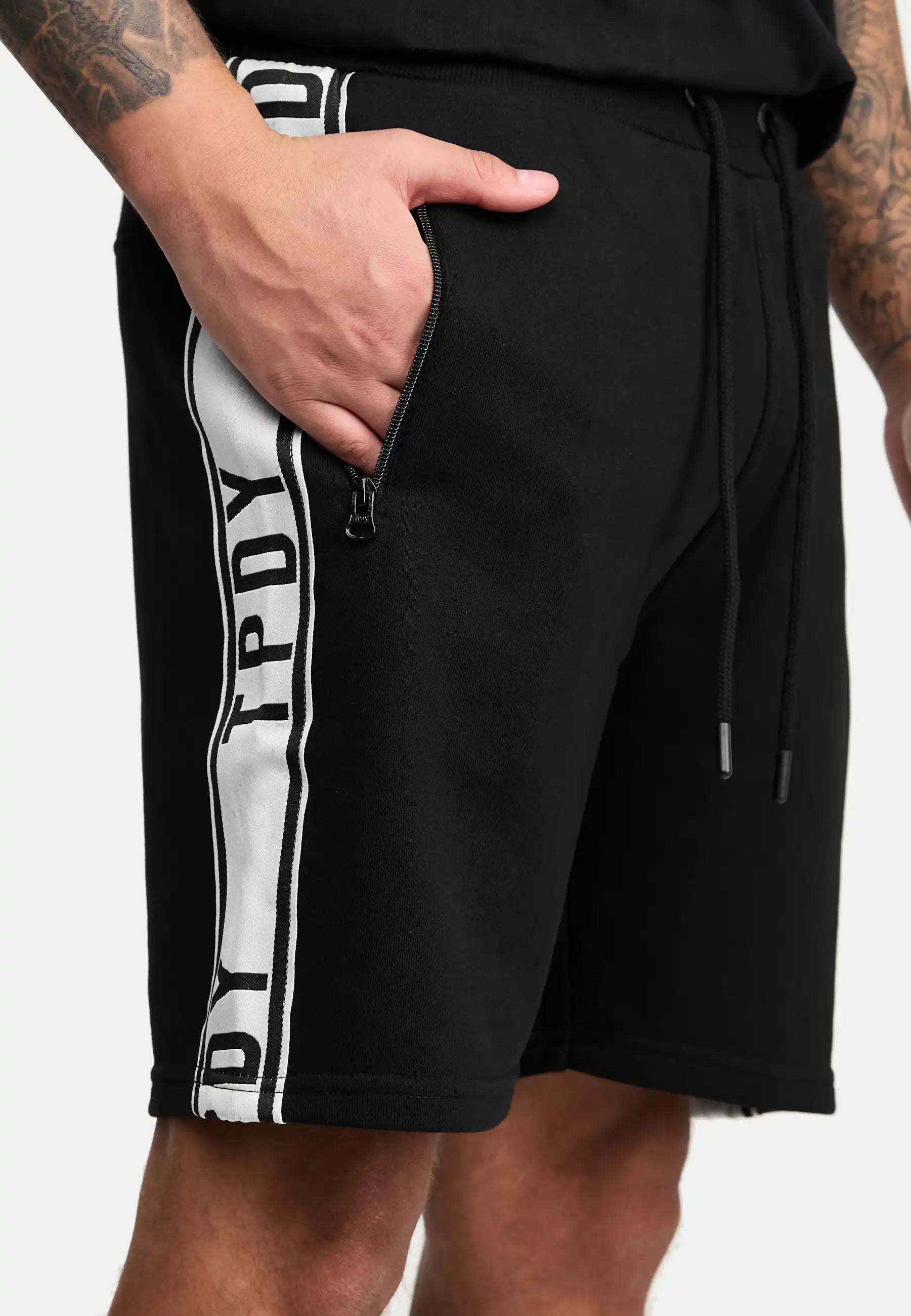 Eine Person trägt trueprodigy Benjamin Logo Shorts mit schwarzem Stoff, einem weißen vertikalen Streifen mit schwarzen Buchstaben und einer Reißverschlusstasche. Nur der untere Teil des Oberkörpers und die Oberschenkel sind sichtbar.