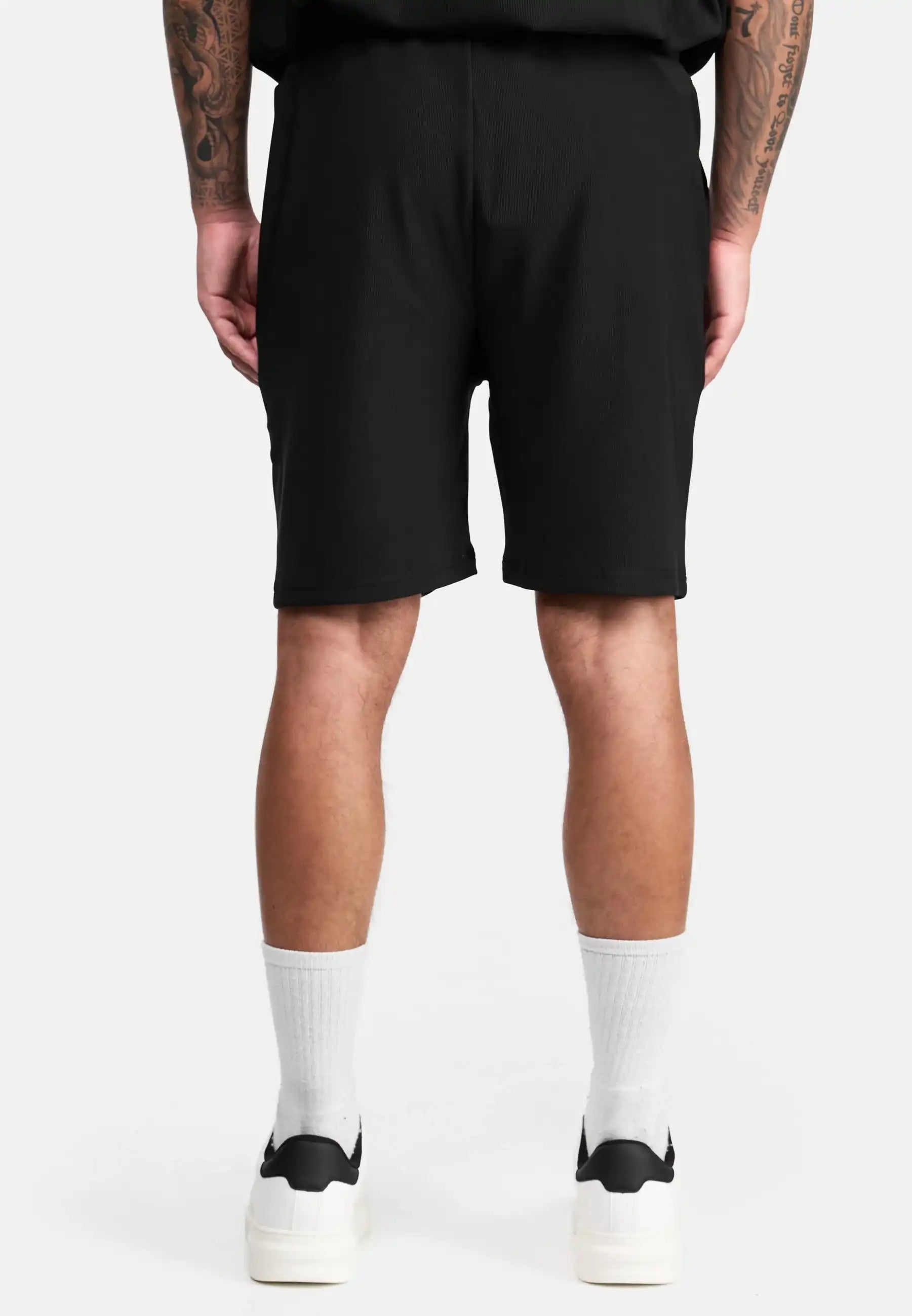Eine Person steht mit dem Rücken zur Kamera, trägt die schwarze trueprodigy Samuel Short mit Stickerei, ein schwarzes Shirt, weiße Crew-Socken und schwarze trueprodigy Sneaker. Ihre tätowierten Arme hängen entspannt herab vor weißem Hintergrund.