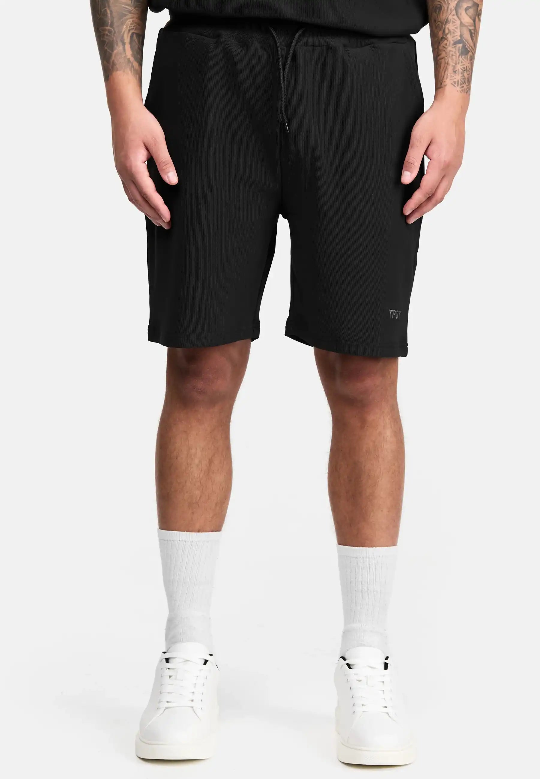 Eine Person trägt die Samuel Short mit Stickerei von trueprodigy in Schwarz und ein schwarzes Shirt, dazu weiße Crew-Socken und Sneakers. Die Arme hängen locker an den Seiten, das Foto zeigt sie von den Schultern abwärts.