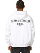 Eine Person steht mit dem Rücken zur Kamera vor einem weißen Hintergrund und trägt den übergroßen Unisex-Hoodie Aaren von trueprodigy. Dieser weiße Hoodie trägt in kräftigem Schwarz den Aufdruck „INTREPID FEARLESS TRUE PRODIGY“ und wird durch schwarze Jeans ergänzt.