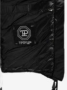 Nahaufnahme der Adan F Steppjacke mit Kapuze von trueprodigy. Sie zeigt eine schwarze gesteppte Innenseite mit einem Patch mit weißen geometrischen Mustern und dem Text „TPDY//®“. Die teilweise geöffnete Jacke zeigt ihr wasserabweisendes Innenfutter.