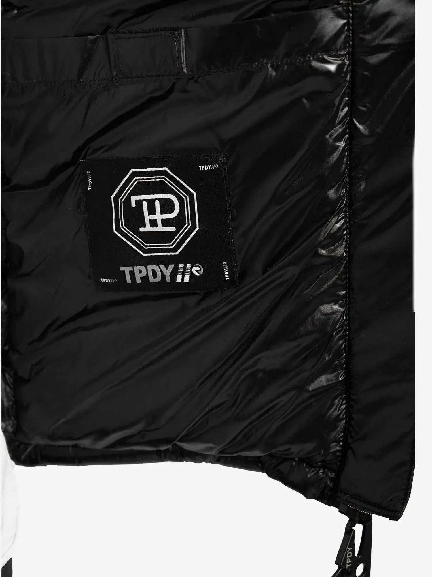 Nahaufnahme der Adan F Steppjacke mit Kapuze von trueprodigy. Sie zeigt eine schwarze gesteppte Innenseite mit einem Patch mit weißen geometrischen Mustern und dem Text „TPDY//®“. Die teilweise geöffnete Jacke zeigt ihr wasserabweisendes Innenfutter.