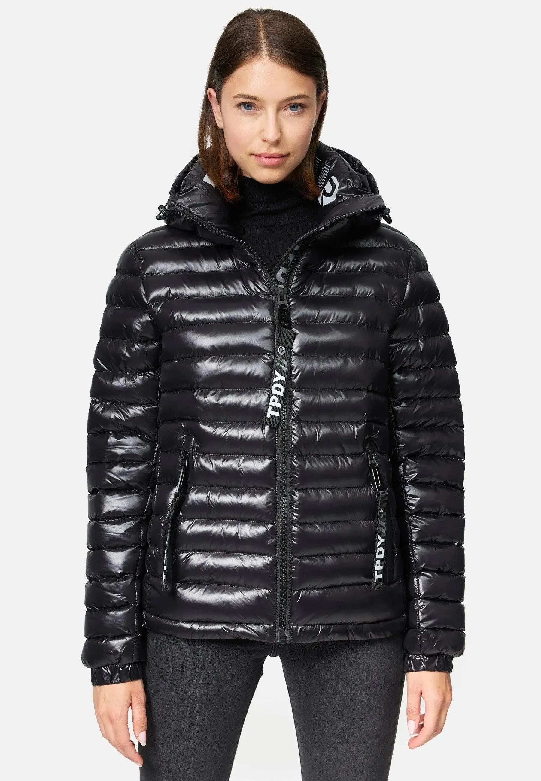 Eine Person in einer glänzend schwarzen Trueprodigy Adan F Steppjacke mit Kapuze steht vor einem schlichten weißen Hintergrund. Die wasserabweisende Jacke hat sichtbare Reißverschlüsse, und die Person trägt außerdem dunkle Jeans und hat langes dunkles Haar.