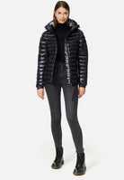 Eine Frau trägt die Adan F Steppjacke mit Kapuze von trueprodigy – eine glänzende, stylische wasserabweisende schwarze Steppjacke. Dazu trägt sie einen schwarzen Rollkragenpullover, dunkelgraue Röhrenjeans und schwarze Schnürstiefel. Ihr glattes braunes Haar umrahmt ihr leicht lächelndes Gesicht vor einem weißen Hintergrund.
