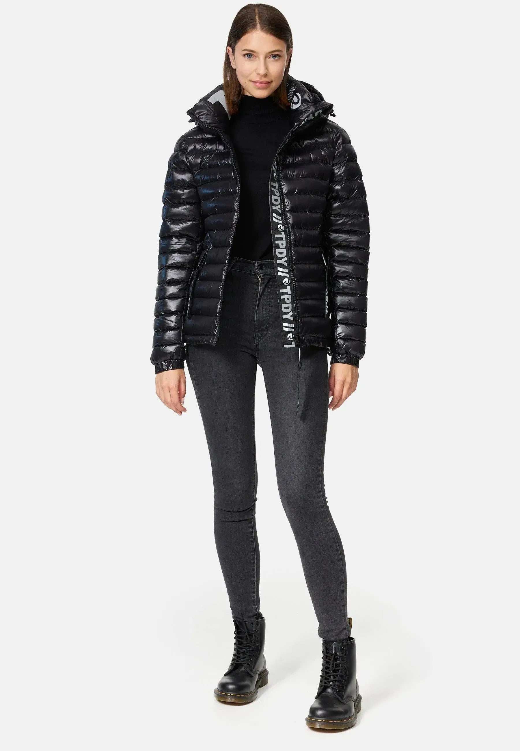 Eine Frau trägt die Adan F Steppjacke mit Kapuze von trueprodigy – eine glänzende, stylische wasserabweisende schwarze Steppjacke. Dazu trägt sie einen schwarzen Rollkragenpullover, dunkelgraue Röhrenjeans und schwarze Schnürstiefel. Ihr glattes braunes Haar umrahmt ihr leicht lächelndes Gesicht vor einem weißen Hintergrund.