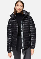 Die Person trägt eine Steppjacke mit Kapuze von trueprodigy Adan F über einem schwarzen Oberteil und dunklen Jeans. Der Reißverschluss der wasserabweisenden schwarzen Jacke ist leicht geöffnet, sodass auf dem Reißverschluss ein Logo zu sehen ist. Sie hat schulterlanges braunes Haar und steht vor einem schlichten weißen Hintergrund.