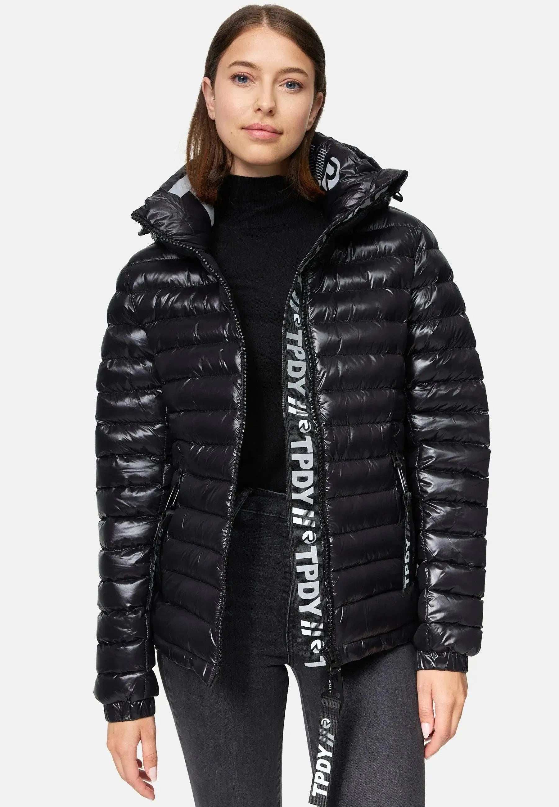 Die Person trägt eine Steppjacke mit Kapuze von trueprodigy Adan F über einem schwarzen Oberteil und dunklen Jeans. Der Reißverschluss der wasserabweisenden schwarzen Jacke ist leicht geöffnet, sodass auf dem Reißverschluss ein Logo zu sehen ist. Sie hat schulterlanges braunes Haar und steht vor einem schlichten weißen Hintergrund.