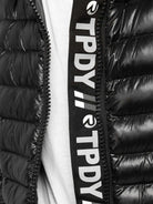 Eine Nahaufnahme der Adan F Steppjacke mit Kapuze in Schwarz von trueprodigy zeigt ihr gestepptes Design mit sichtbarem Reißverschluss. Der Reißverschluss hat eine schwarze Stoffauflage mit vertikalem weißen „TPDY“-Schriftzug, der auf den Stil der Marke hinweist. Darunter lugt ein weißes T-Shirt hervor.