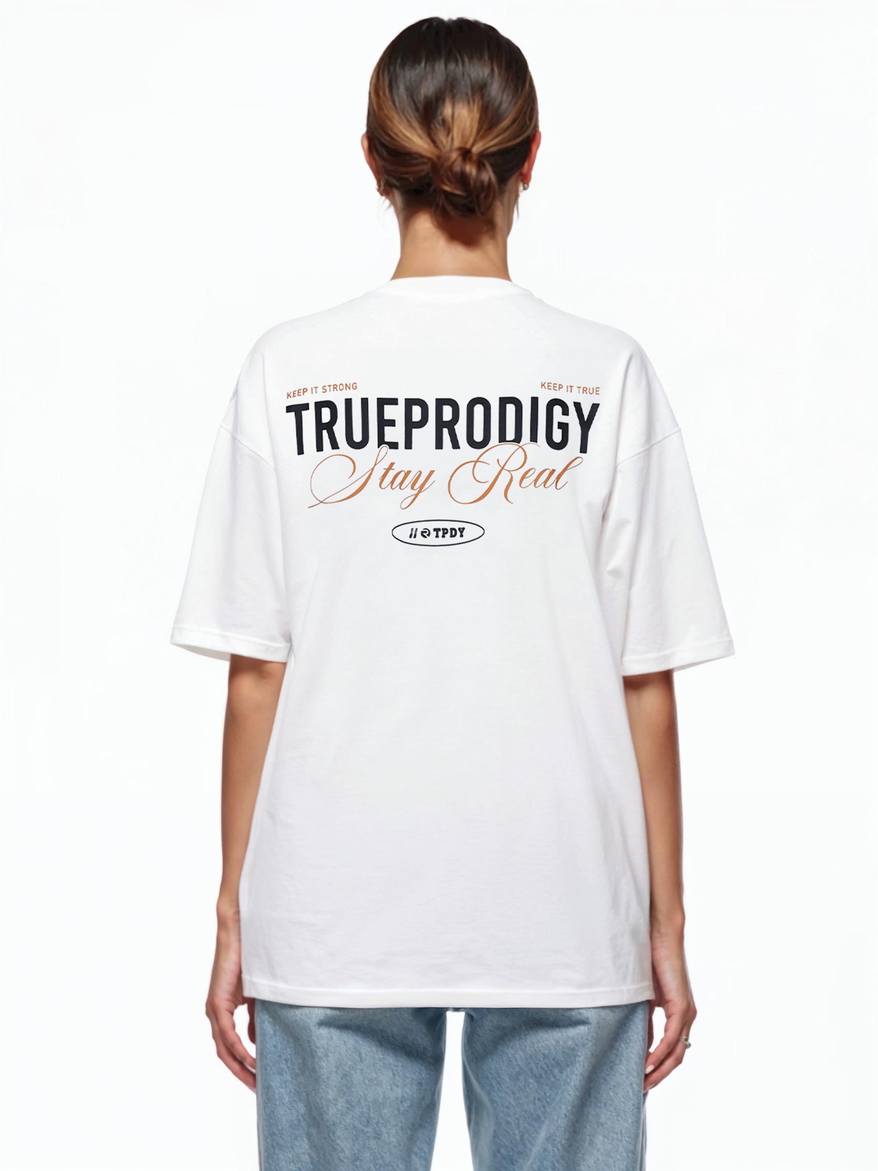 Eine Person trägt ein weißes T-Shirt mit dem Aufdruck „TRUEPRODIGY Stay Real“ in schwarz-orangefarbener Schrift auf der Rückseite. Das T-Shirt enthält Hashtags und zusätzlichen kleineren Text. Die Person hat mittellanges Haar und trägt Blue Jeans. Der Blick ist von der Kamera abgewandt.