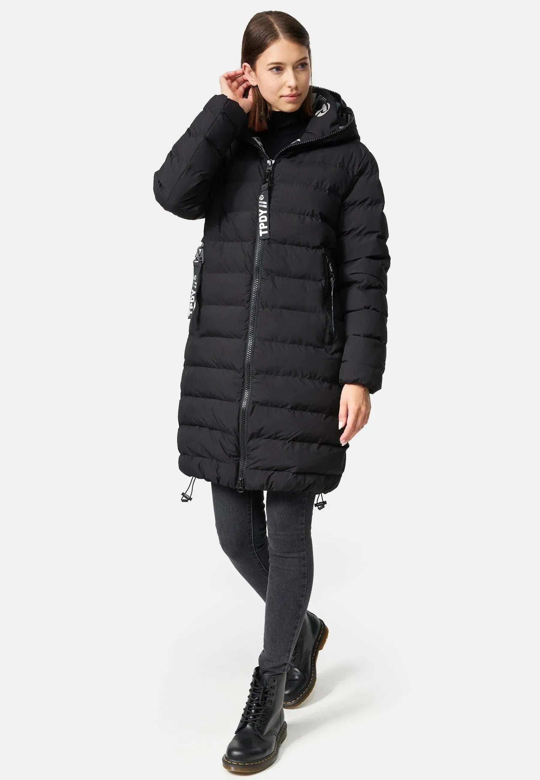 Eine Person, die eine Chester F Lange Pufferjacke mit Logo von trueprodigy trägt, dazu schwarze Jeans und Stiefel, steht vor einem schlichten Hintergrund. Eine Hand ist in der Nähe ihres Kopfes erhoben, während die andere an ihrer Seite ruht. Die schwarze Jacke verfügt über einen stylischen Reißverschluss und eine Kapuze.