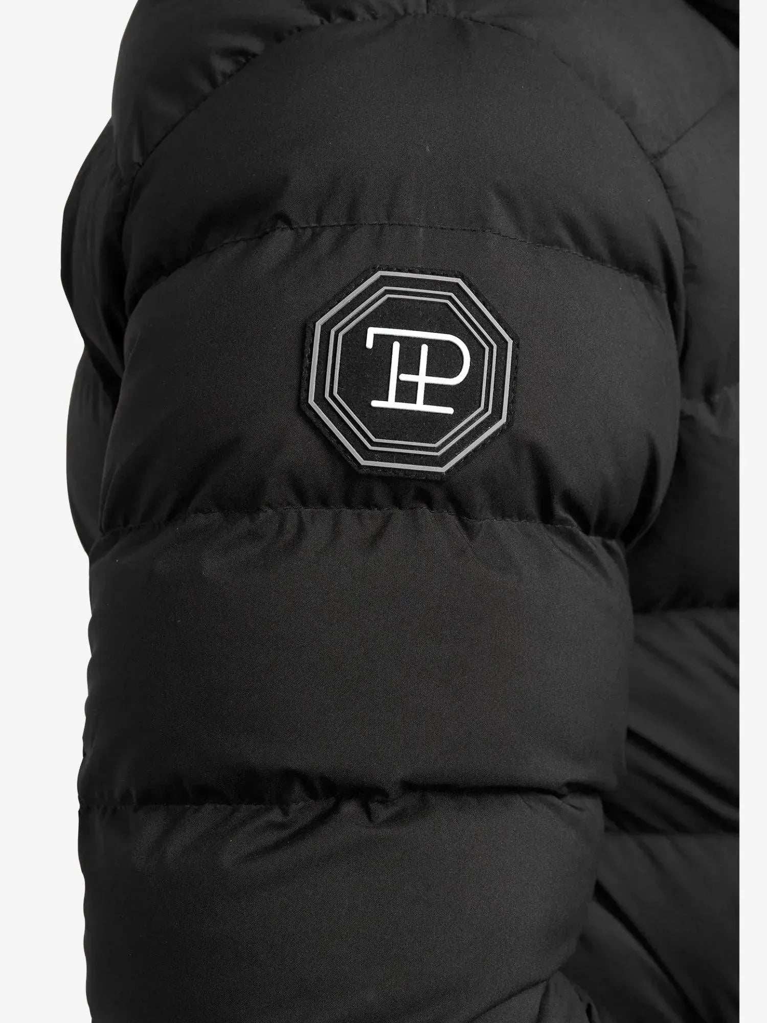 Nahaufnahme eines Ärmels einer Chester F Lange Pufferjacke von trueprodigy, mit einem schwarzen achteckigen Patch mit ineinandergreifenden weißen Buchstaben. Der gesteppte Stoff sorgt für Wärmeisolierung, während das wasserabweisende Material und der Reißverschluss bei Nässe für Funktionalität sorgen.