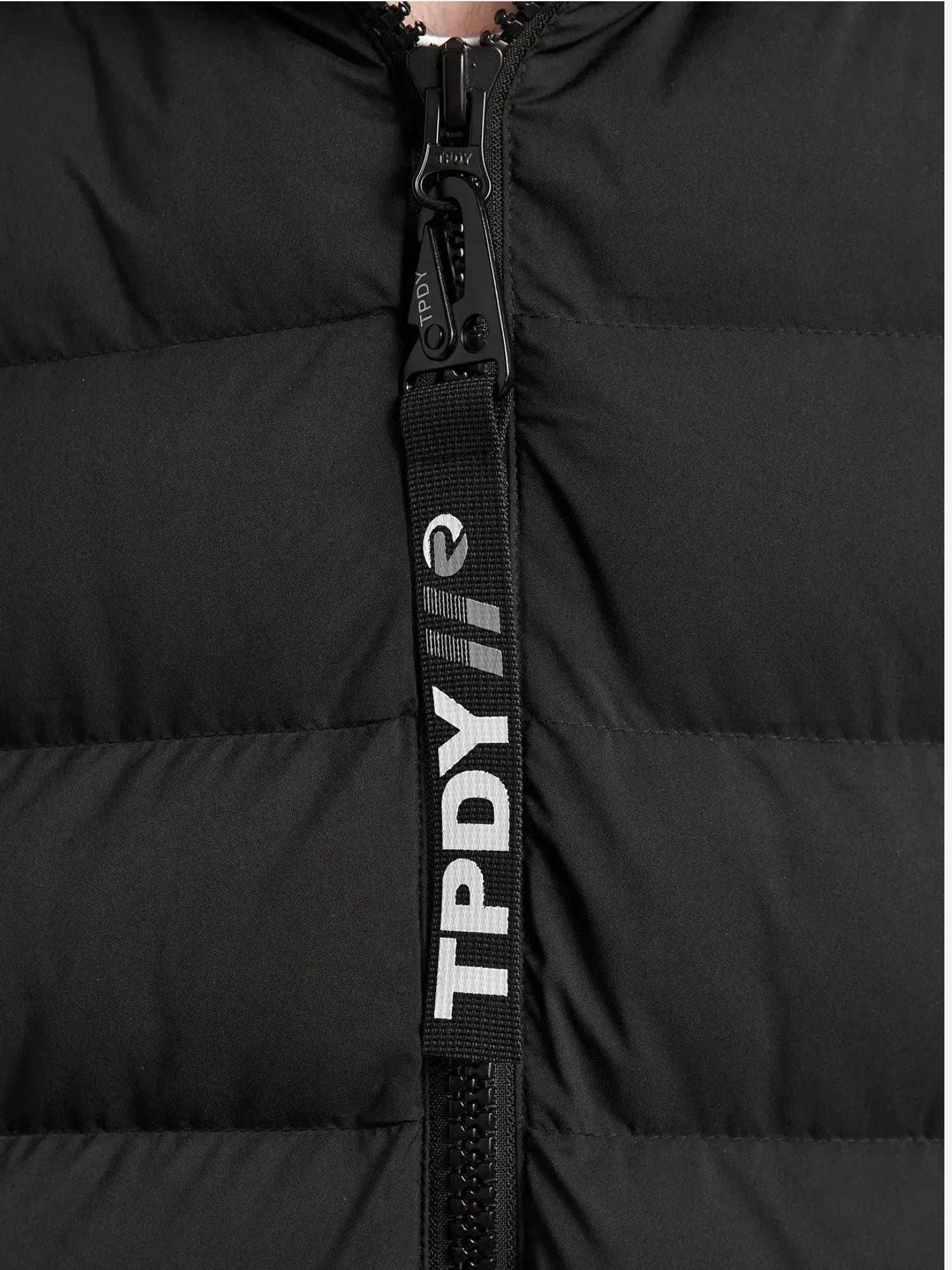 Nahaufnahme einer schwarzen gesteppten Chester F Lange Pufferjacke mit Logo von trueprodigy, mit Reißverschluss mit schwarzer Lasche, auf der „TPDY“ in Weiß steht, und einem Logo mit diagonalem Streifen. Der gepolsterte Stoff ist ein wasserabweisendes Material, das Stil und Schutz vereint.