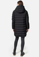 Eine Figur steht mit abgewandtem Gesicht in einer Chester F Lange Pufferjacke mit Logo von trueprodigy. Die lange schwarze Pufferjacke besteht aus wasserabweisendem Material und hat einen Reißverschluss, der Licht einfängt. Sie trägt sie mit dunkelgrauen Röhrenjeans und schwarzen Stiefeletten auf einem schlichten weißen Hintergrund.