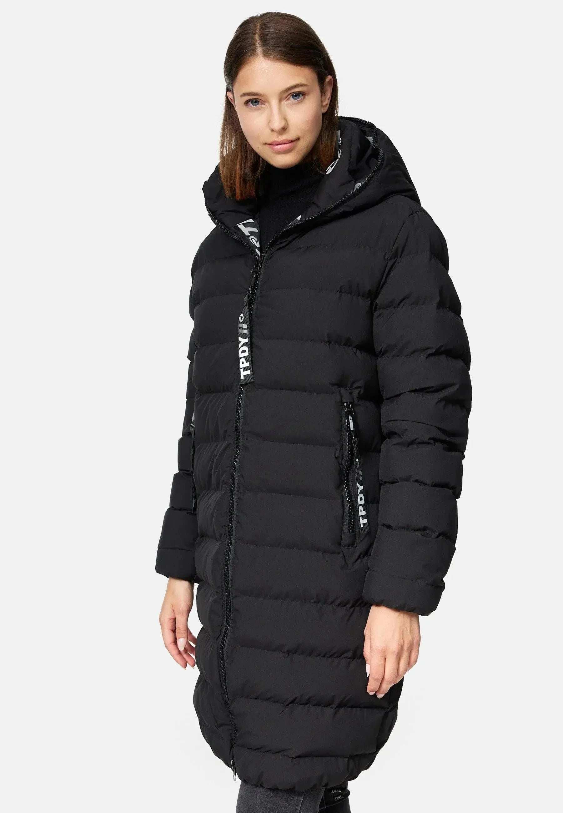 Eine Person mit schulterlangem braunem Haar posiert mühelos vor einem weißen Hintergrund und trägt eine Chester F Lange Pufferjacke mit Logo von trueprodigy. Der lange schwarze Parka ist aus wasserabweisendem Material gefertigt und verfügt über eine Kapuze, vertikale Reißverschlusstaschen und einen Markenreißverschluss.