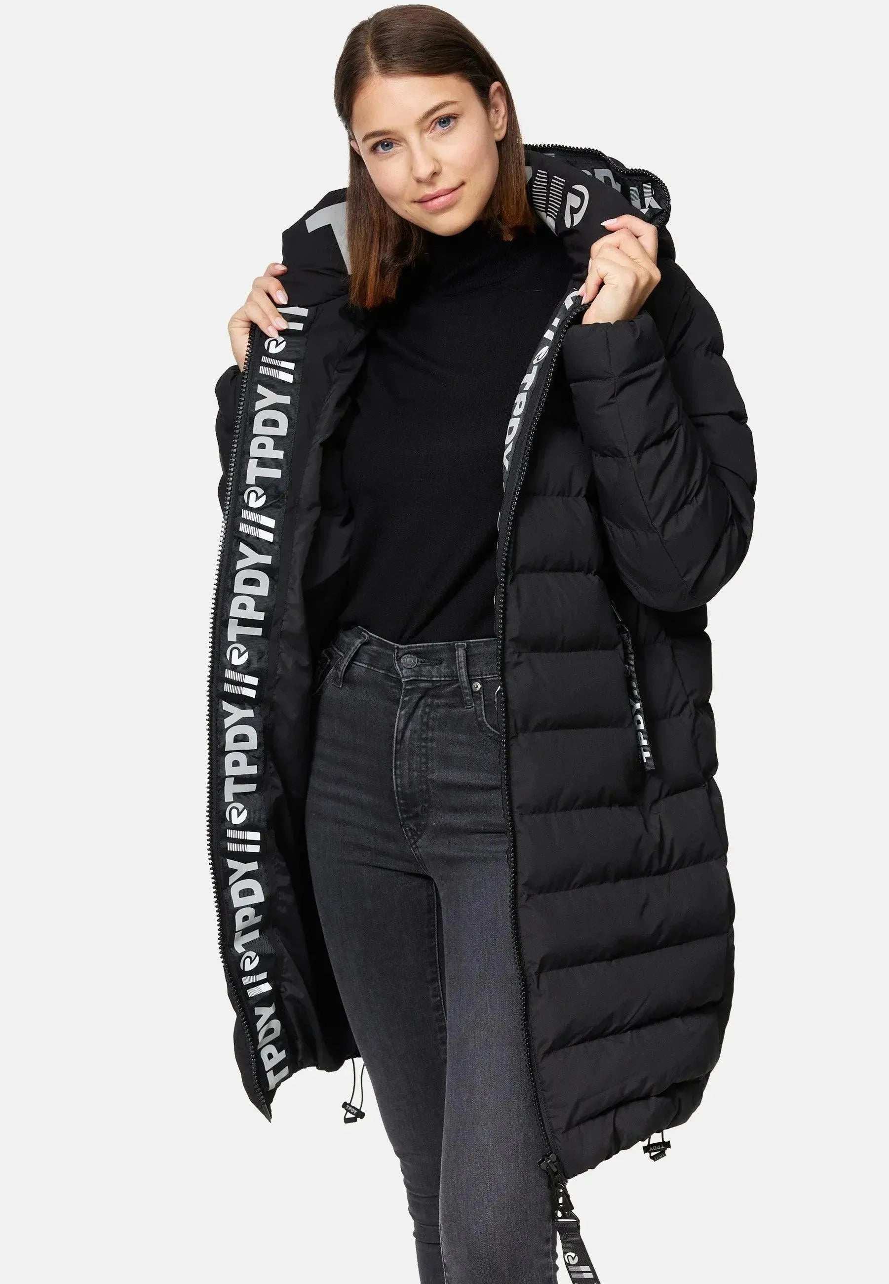 Eine Frau in schwarzem Rollkragenpullover und Jeans trägt die Chester F Lange Pufferjacke mit Logo von trueprodigy, gefertigt aus wasserabweisendem Material. Die Pufferjacke mit teilweisem Reißverschluss hat ein Futter mit „PDVI“-Muster. Mit schulterlangem Haar blickt sie selbstbewusst in die Kamera.