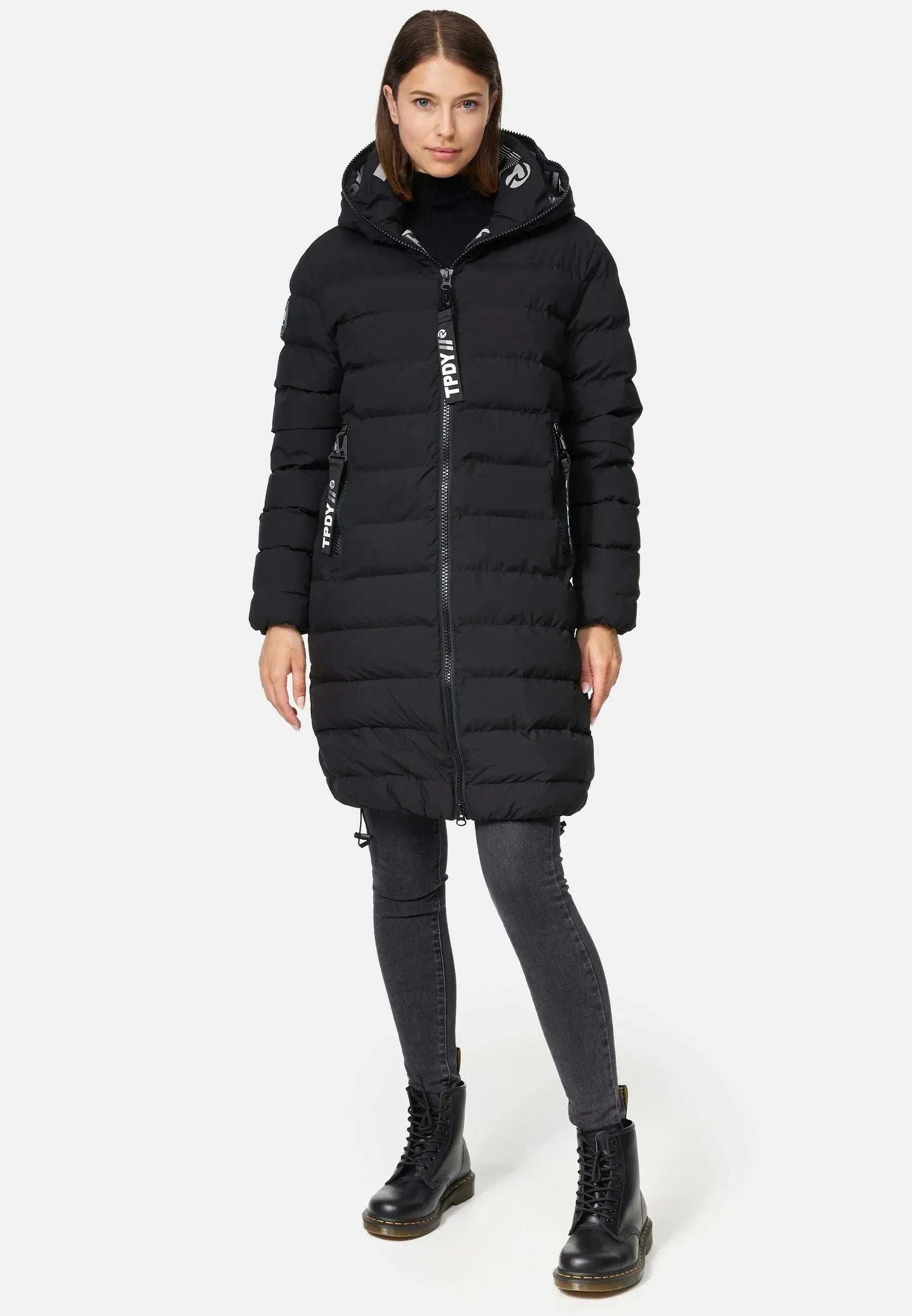 Eine Frau steht vor einem schlichten Hintergrund und trägt eine lange schwarze Trueprodigy Chester F Lange Pufferjacke mit Kapuze. Sie ist aus wasserabweisendem Material gefertigt und verfügt über einen eleganten Reißverschluss. Sie kombiniert sie mit dunklen Jeans und schwarzen Stiefeln, ihr glattes Haar ist offen, während sie in die Kamera blickt.