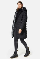 Eine Frau trägt eine lange schwarze Chester F Lange Pufferjacke mit Logo von trueprodigy, gefertigt aus wasserabweisendem Material. Kombiniert mit schwarzen Röhrenjeans und Stiefeln zeigt der Mantel ein vertikales Logo entlang des Reißverschlusses. Sie steht vor einem schlichten weißen Hintergrund.