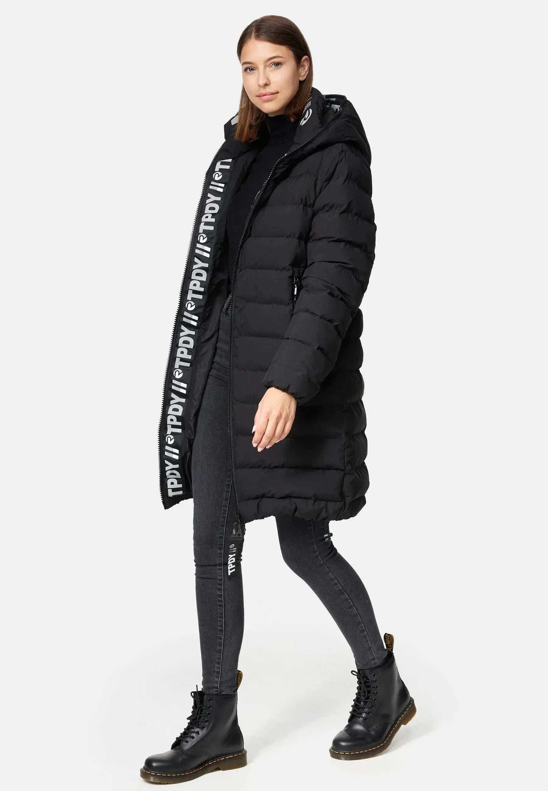 Eine Frau trägt eine lange schwarze Chester F Lange Pufferjacke mit Logo von trueprodigy, gefertigt aus wasserabweisendem Material. Kombiniert mit schwarzen Röhrenjeans und Stiefeln zeigt der Mantel ein vertikales Logo entlang des Reißverschlusses. Sie steht vor einem schlichten weißen Hintergrund.