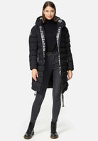 Eine Person präsentiert die Chester F Lange Pufferjacke mit Logo von trueprodigy, einen schwarzen Kapuzenmantel aus wasserabweisendem Material. Er hat einen weißen Text auf der Innenseite, schwarze Schnürsenkel und einen eleganten Reißverschluss und wird über einem Rollkragenpullover und Jeans mit schwarzen Stiefeln vor einem schlichten weißen Hintergrund getragen.