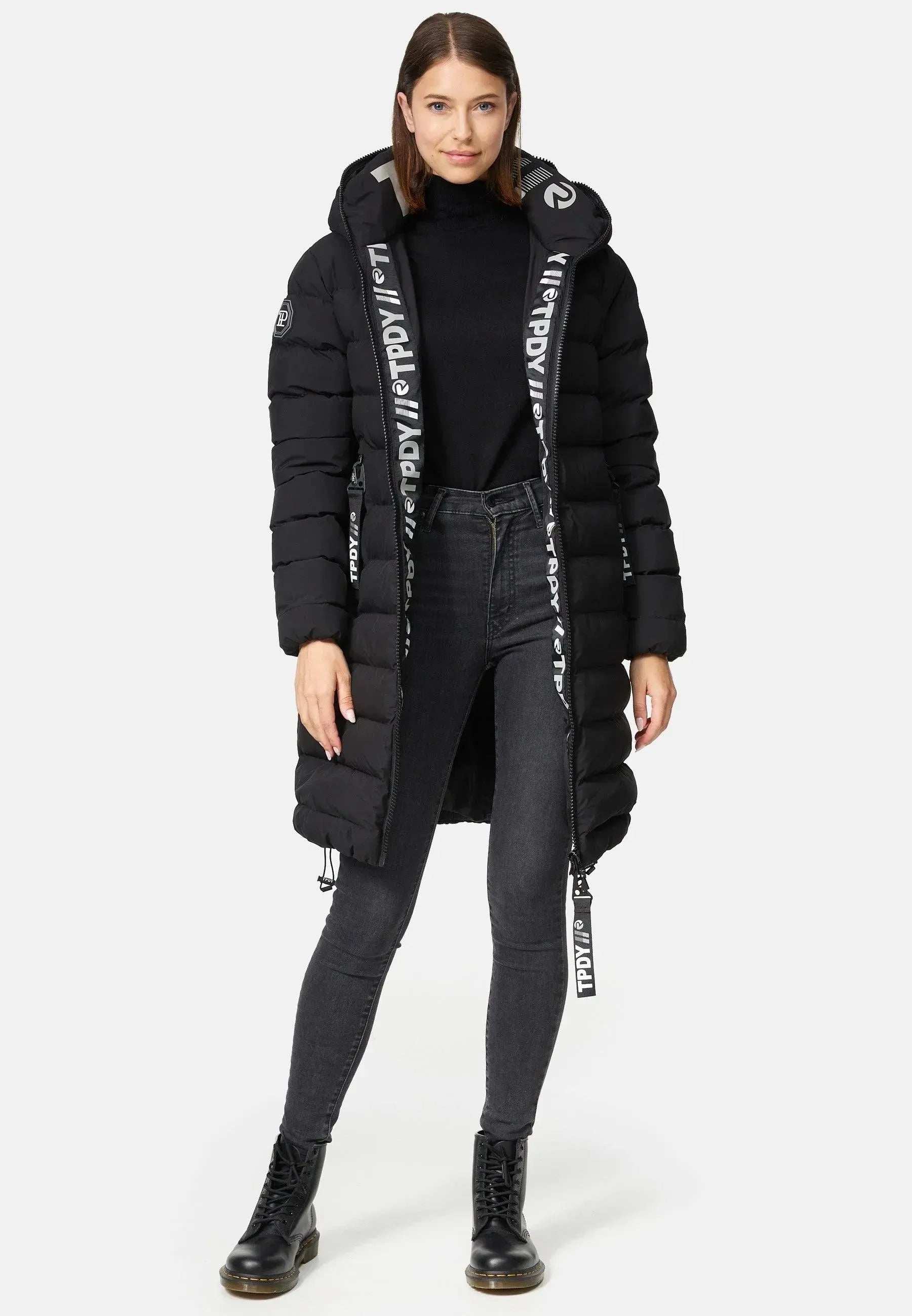 Eine Person präsentiert die Chester F Lange Pufferjacke mit Logo von trueprodigy, einen schwarzen Kapuzenmantel aus wasserabweisendem Material. Er hat einen weißen Text auf der Innenseite, schwarze Schnürsenkel und einen eleganten Reißverschluss und wird über einem Rollkragenpullover und Jeans mit schwarzen Stiefeln vor einem schlichten weißen Hintergrund getragen.