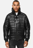 Jemand trägt eine glänzend schwarze Corey Steppjacke mit Stehkragen von trueprodigy mit „TPDW“ auf der Vorderseite und den Ärmeln. Sie kombinieren sie mit dunklen Hosen auf einem strahlend weißen Hintergrund.